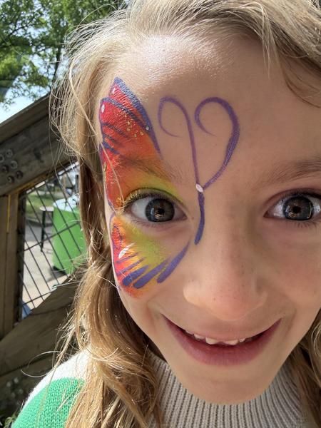 A young child with a vibrant butterfly painted on their face smiles brightly at the camera